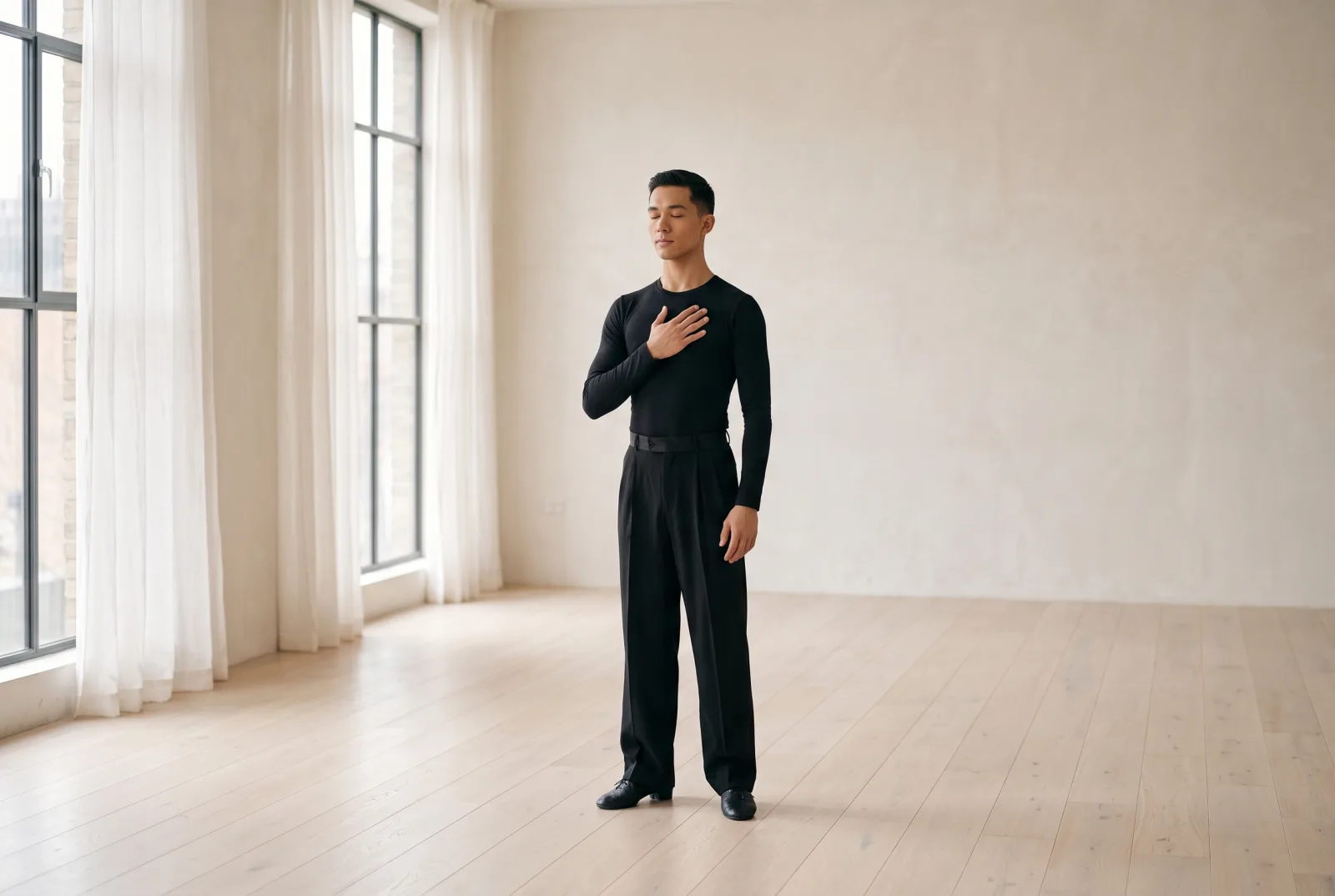 A dancer standing still in an empty studio, eyes closed, one hand on his chest.
